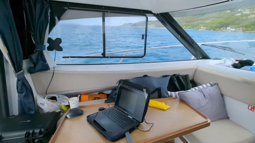 A laptop computer on a table inside a small boat off the coast.