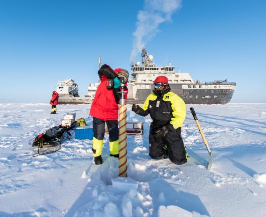Researchers drilling a hole in the polar ice with the research icebreaker RV Kronprins Haakon in the background.
