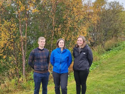 Three people standing outdoors in front of trees with autumn foliage.