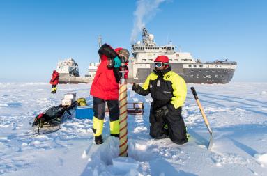Researchers drilling a hole in the polar ice with the research icebreaker RV Kronprins Haakon in the background.