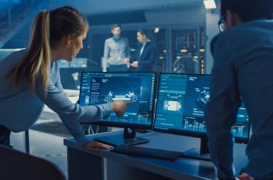 A woman in a high-tech work setting points at a computer screen to show a colleague something. 